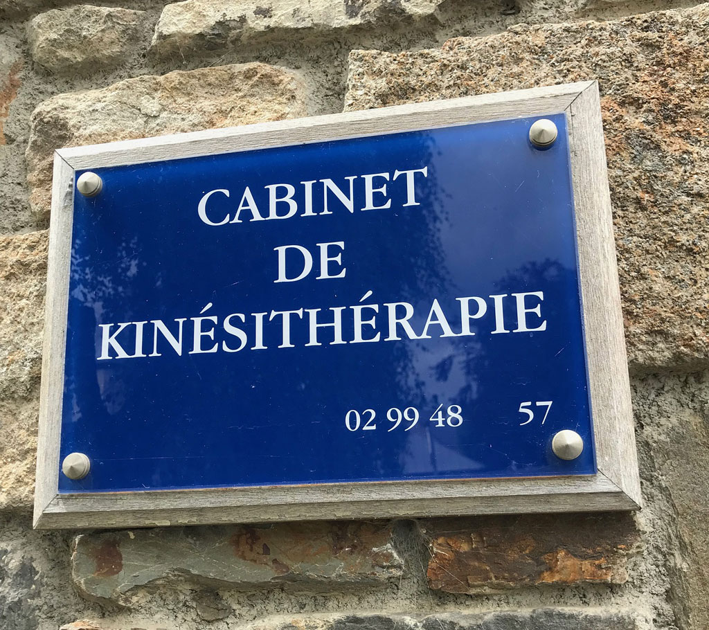 Cette image montre une plaque professionnelle rectangulaire en métal argenté. La surface de la plaque est recouverte d'une peinture lisse et brillante qui reflète la lumière du soleil. Sur le dessus se trouve un logo, des lettres en relief et une inscription noire qui indique le nom de l'entreprise et son adresse. Les bords sont décorés par des ornements moulés à la main qui ajoutent une touche artistique à cette plaque digne de confiance.