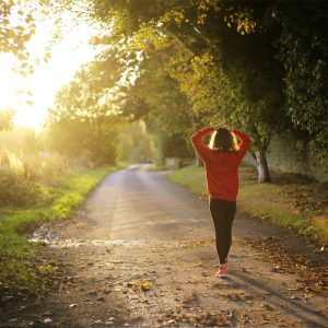 Cette photo représente une personne en train de pratiquer un footing. Elle est vêtue d'un survêtement bleu et porte des chaussures de course blanches, elle court avec détermination sur un chemin qui s'étend à l'horizon. La nature luxuriante et verdoyante l'entoure, avec des arbres aux branches touffues offrant une ombre bienfaisante. On peut imaginer que cette activité physique sert à libérer le stress post-traumatique, ce qui est renforcé par la sensation de liberté que donne la vaste étendue devant elle.