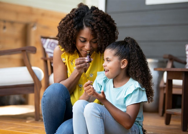 Une femme et une jeune fille souriantes assises sur une terrasse en bois. La femme, vêtue d'un haut jaune, tient un petit flacon d'huile essentielle près de son visage tandis que l'enfant la regarde joyeusement.