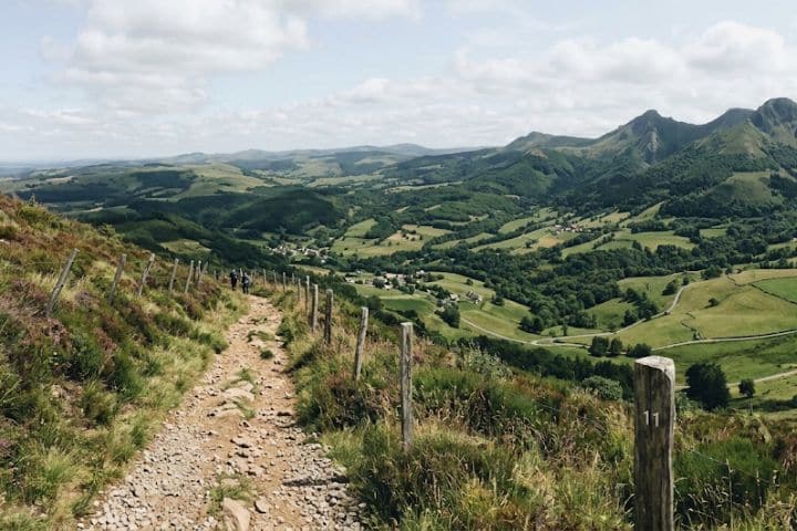 Sentier de randonnée caillouteux bordé de piquets en bois, avec vue sur une vallée verdoyante et des sommets sous un ciel nuageux